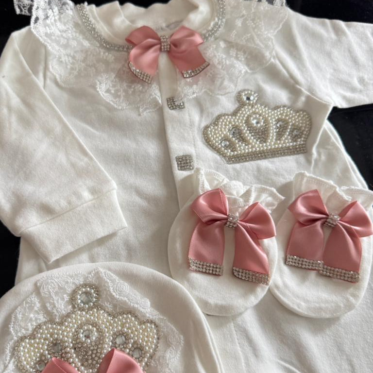 A white baby girl's royal-themed outfit set including a long-sleeve top, booties, a bonnet, and a matching bib, decorated with pink satin bows, lace, rhinestone-embellished crowns, and pearl details.
