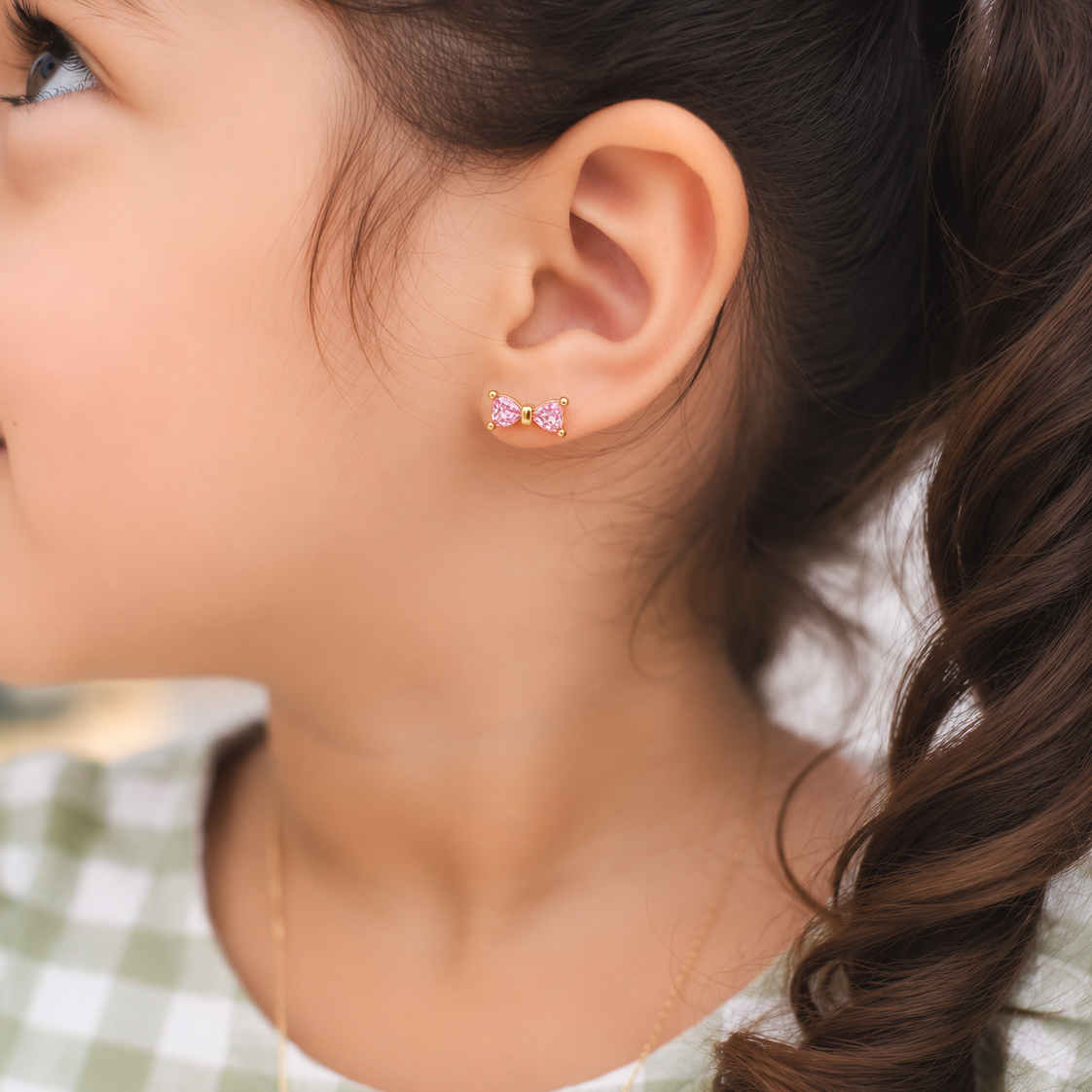 A close-up of a young girl's ear wearing a small pink bow stud earring, with her brown hair in a braid and a green and white checkered top visible.