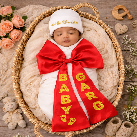 Newborn baby wrapped in a red sash with 'Baby Girl' and a white hat with 'Hello World' text, surrounded by flowers and toys on a wooden floor.
