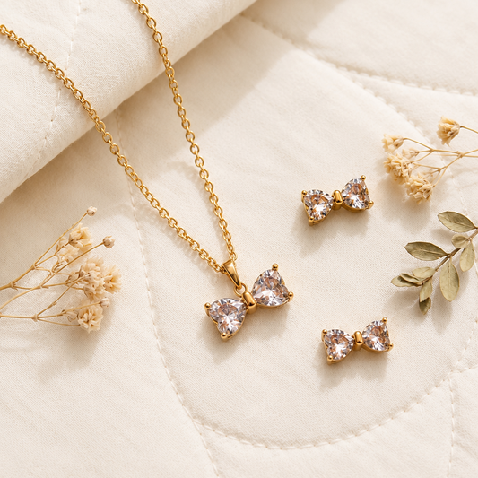 Gold-tone necklace and matching stud earrings with small bow-shaped clear gemstone pendants, arranged on a cream quilted fabric with dried floral props.