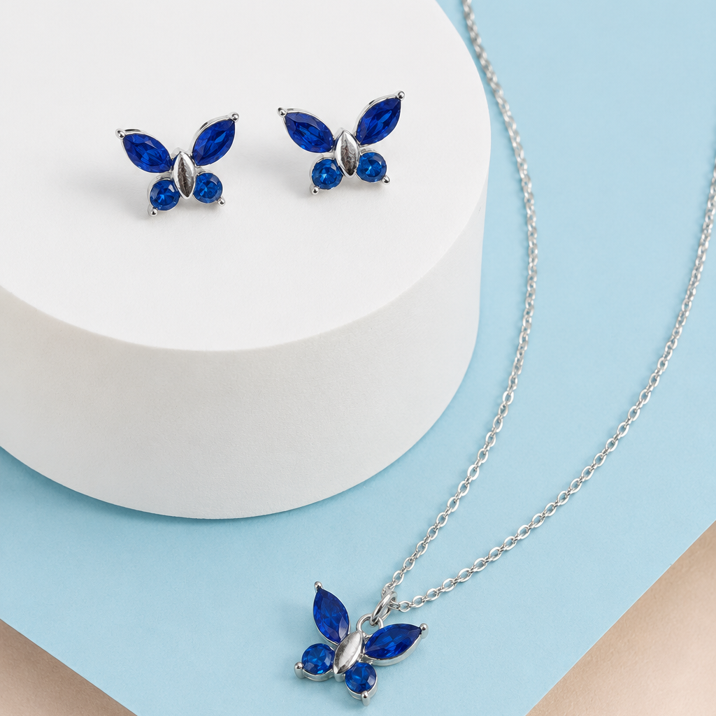 Silver butterfly jewelry set with deep blue stones, including a necklace with a matching pendant and a pair of stud earrings, displayed on a white cylindrical pedestal and a light blue background.
