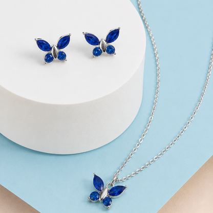 Silver butterfly jewelry set with deep blue stones, including a necklace with a matching pendant and a pair of stud earrings, displayed on a white cylindrical pedestal and a light blue background.