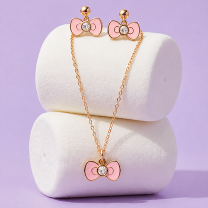 A gold-tone jewelry set featuring a necklace and matching stud earrings, both decorated with pink bow charms and a clear rhinestone in the center, displayed on white cylindrical stands against a lavender background.