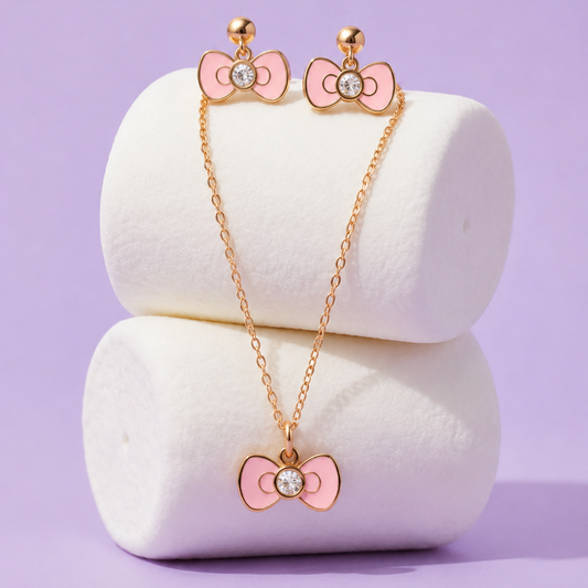 A gold-tone jewelry set featuring a necklace and matching stud earrings, both decorated with pink bow charms and a clear rhinestone in the center, displayed on white cylindrical stands against a lavender background.