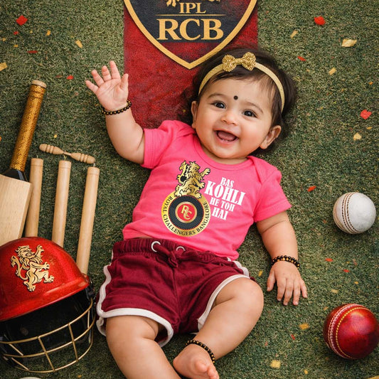 Baby wearing a pink shirt with RCB logo surrounded by cricket equipment on a green background