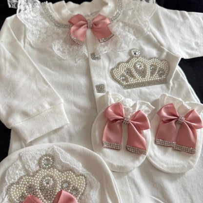 A white baby girl's royal-themed outfit set including a long-sleeve top, booties, a bonnet, and a matching bib, decorated with pink satin bows, lace, rhinestone-embellished crowns, and pearl details.