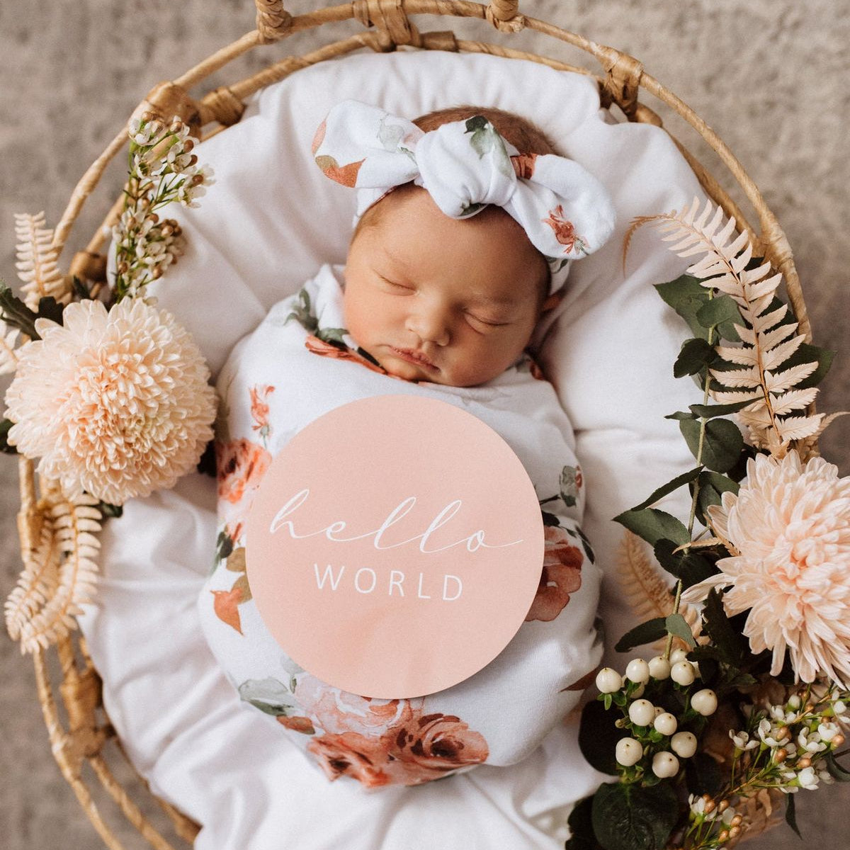 Newborn baby in a basket with floral decorations and a 'hello world' sign.