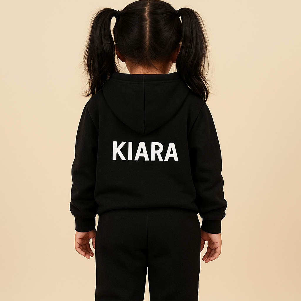 Child wearing a black hoodie and pants with 'KIARA' printed on the back, standing against a beige wall.