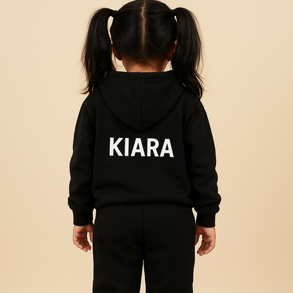 Child wearing a black hoodie and pants with 'KIARA' printed on the back, standing against a beige wall.