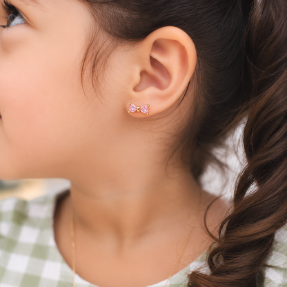 A close-up of a young girl's ear wearing a small pink bow stud earring, with her brown hair in a braid and a green and white checkered top visible.