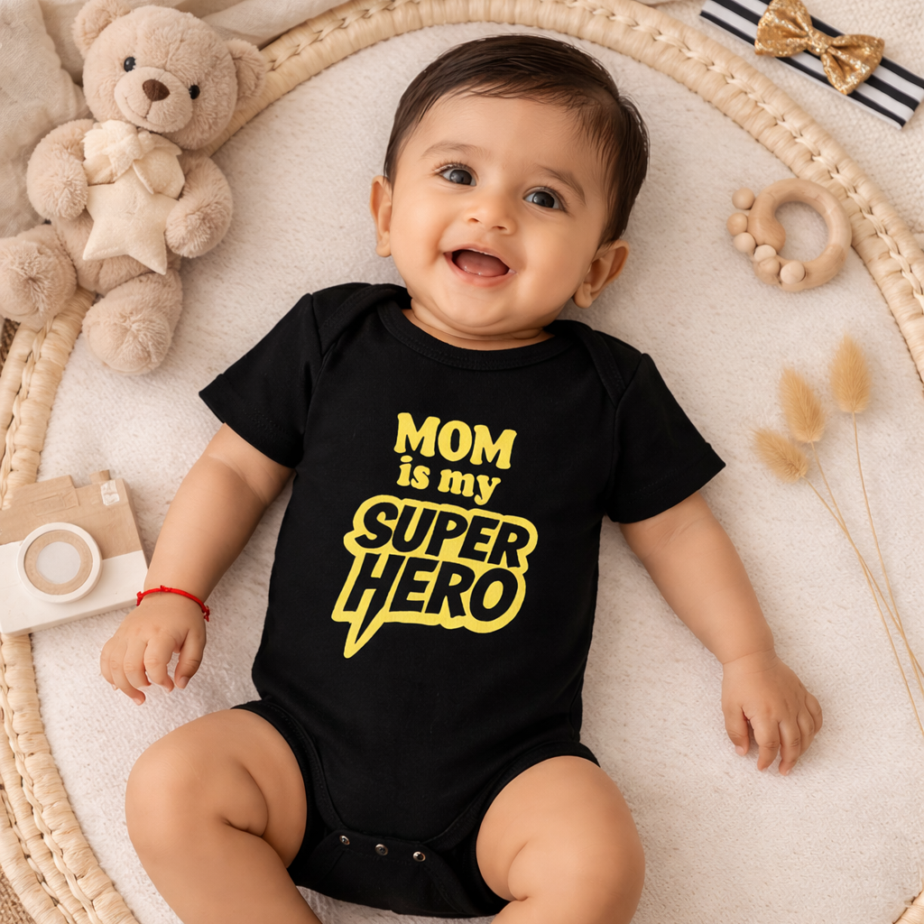 Baby wearing a black onesie with 'Mom is my super hero' text, surrounded by toys on a textured surface.