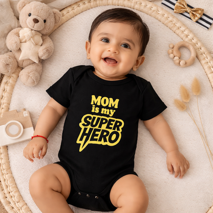Baby wearing a black onesie with 'Mom is my super hero' text, surrounded by toys on a textured surface.