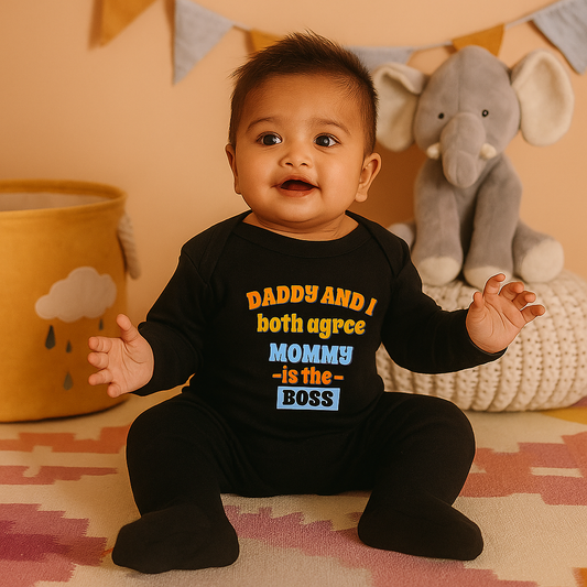 Baby wearing a black onesie with colorful text, sitting on a patterned rug with toys in the background.