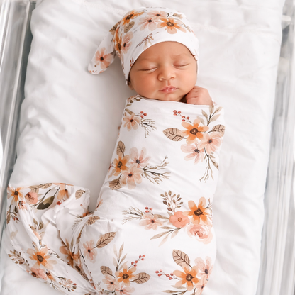 Newborn baby wrapped in a floral swaddle and headband, lying on a white blanket.