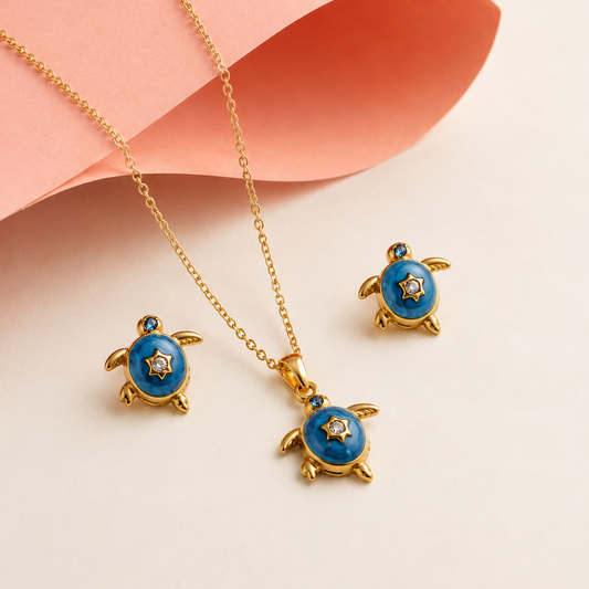 Gold-tone jewelry set featuring a necklace and two matching earrings with sea turtle pendants, decorated with vibrant blue enamel and small white star details, styled on a light beige background with a peach paper accent.